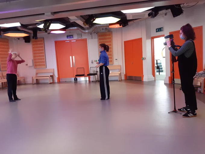 A woman in a pink top and black trousers dancers, her hands held by her face. The choreographer, another woman watches as another records the movement with a camera. They are in a dance studio with bright orange doors.