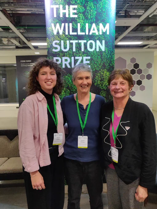 Photo of Pride of Place members Naomi, Susan and Jane smiling and standing in front of a William Sutton Prize banner 