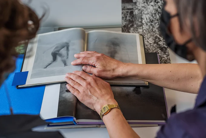  Two people in an archive are looking at the open spread of pages of a photography book. One person is holding the book up in the air. They are wearing masks. 