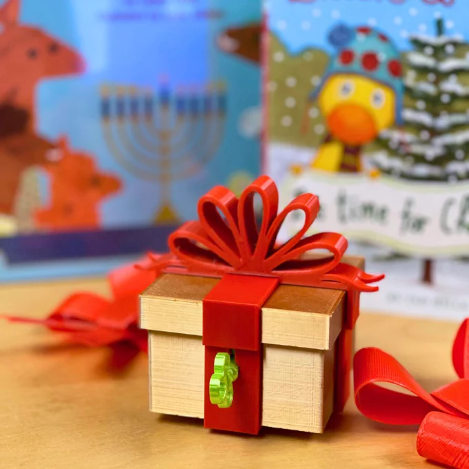 A 3D-printed gift box wrapped in a red 3D-printed bow. A small green key extends from a lock in the front. The box is flanked by two out-of-focus holiday-themed children's books.