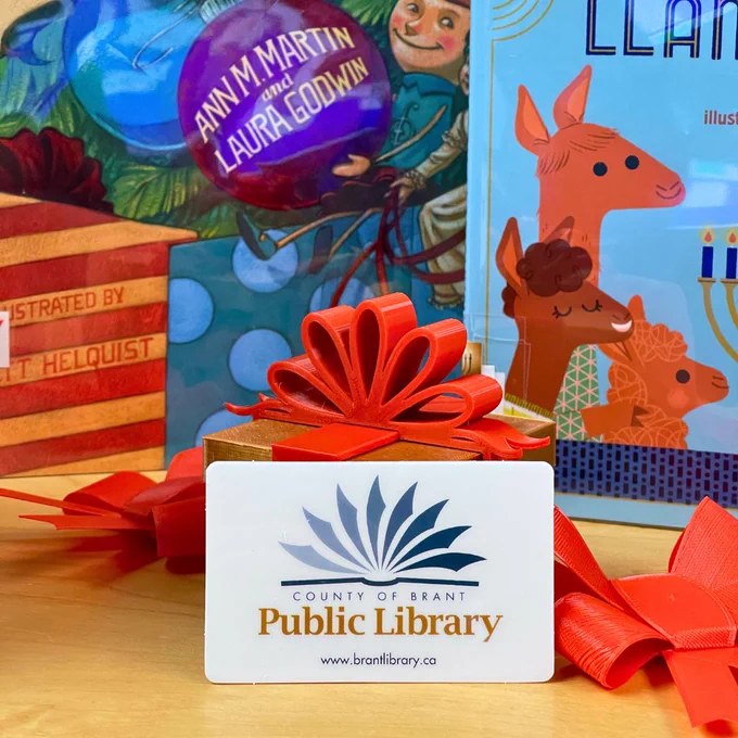 A library card is propped up in front of a 3D printed gift box. Two children's holiday books stand upright in the background.