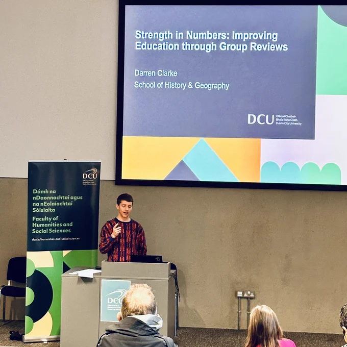 Darren stands at a podium speaking to attendees with a slide behind reading “Strength in Numbers: Improving Education through Group Reviews
Darren Clarke
School of History & Geography”