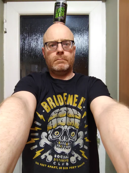 A silly, probably drunken bald man with glasses wearing a black t-shirt for the Brudenell Social Club. Main bit is a skull and crossbones, the top of the skull reads "Stay Home", the bottom text says "six feet apart or six feet under". The main looks mock serious and has a can of beer balanced on his head in similar colour scheme to the tee shirt: orange and white (or light grey) on black.