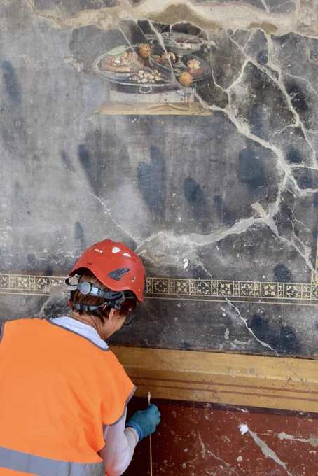 Conservator in high viz jacket and red hard hat patiently cleans the yellow border of the frescoed wall in front of her with a cotton bud. The background of the fresco is black and only interrupted by a depiction of a small scene of a silver laden up with a type of bread covered with dried fruit and a type of sauce; some yellow strawberry tree fruit ; some nuts, and a silver chalice of red wine.