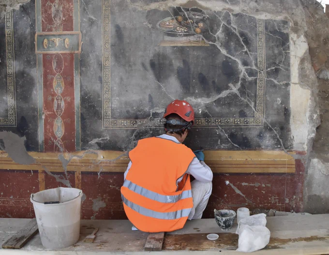 Conservator perched on a wooden plank in high viz jacket and red hard hat patiently cleans the yellow border of the frescoed wall in front of her with a cotton bud. Her tools of her trade lie around her. The background of the fresco is black and only interrupted by a depiction of a small scene of a silver laden up with a type of bread covered with dried fruit and a type of sauce; some yellow strawberry tree fruit ; some nuts, and a silver chalice of red wine. To the left the border is a striking red colour with green edges and has ovals top to tail running down it. There is a small vignette of a bull’s skull framed in golden yellow.