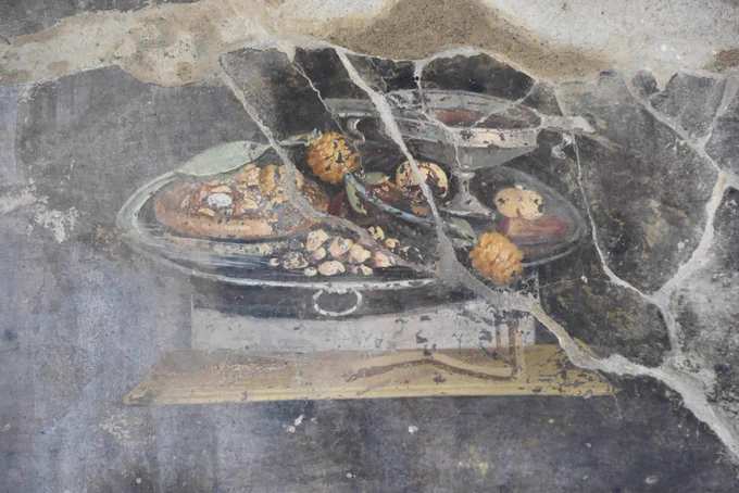 The background of the fresco is black and only interrupted by a depiction of a small scene of a silver laden up with a type of bread covered with dried fruit and a type of sauce; some yellow strawberry tree fruit ; some nuts, and a silver chalice of red wine.