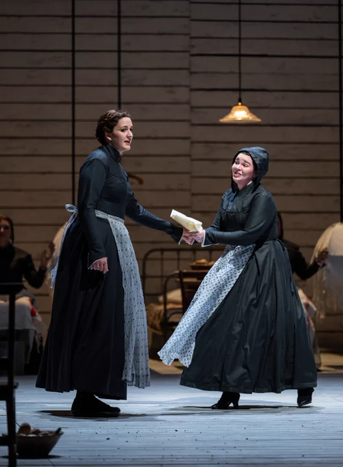 Two women stand in black dresses with light blue patterned aprons. On the left is soprano Lise Davidsen (Jenufa) and on the right is soprano Kathryn Henry (Jana) holding a letter.