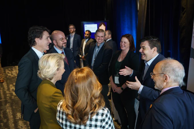 Prime Minister Justin Trudeau, President Charles Michel, and President Ursula von der Leyen are standing and looking at a man, who is standing in front of them. The man is laughing and gesturing with his hands. Other people are standing around them.