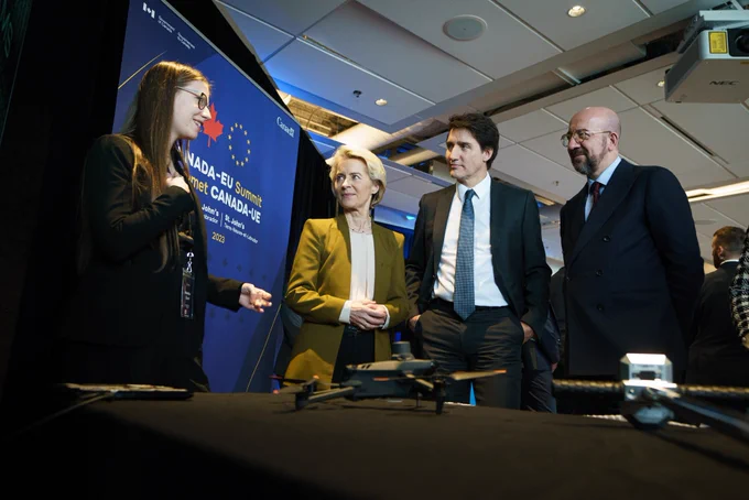 Prime Minister Justin Trudeau, President Charles Michel, and President Ursula von der Leyen are standing and looking at a woman, who is standing in front of them. The woman is speaking and gesturing with her hands. A drone is displayed on the table in front of them.