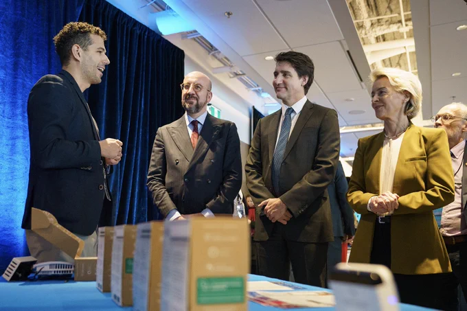 Prime Minister Justin Trudeau, President Charles Michel, and President Ursula von der Leyen are standing and looking at a man, who is standing in front of them and speaking. Other people are standing behind them. Boxes are displayed on the table in front of them.