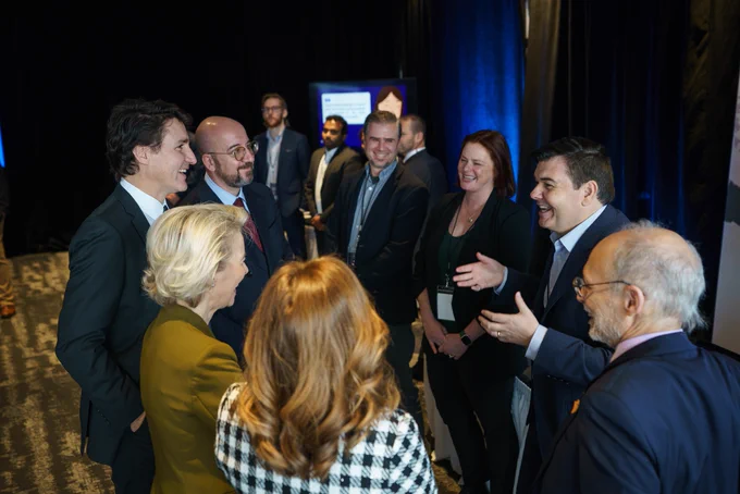 Prime Minister Justin Trudeau, President Charles Michel, and President Ursula von der Leyen are standing and looking at a man, who is standing in front of them. The man is laughing and gesturing with his hands. Other people are standing around them.