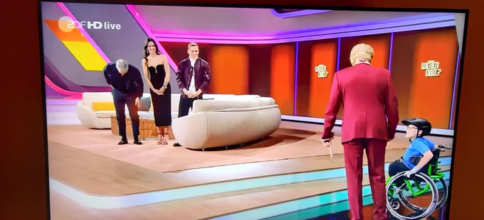 Man sieht die Bühne von Wetten dass. Oben auf der Couch Sebastian Schweinsteiger und Anna Ivanova sowie Matthias Schweighöfer. Unten Thomas Gottschalk mit seinem Jugendlichen Wettgast im Rollstuhl. Die Bühne ist sichtbar nur über zwei Stufen zu erreichen.