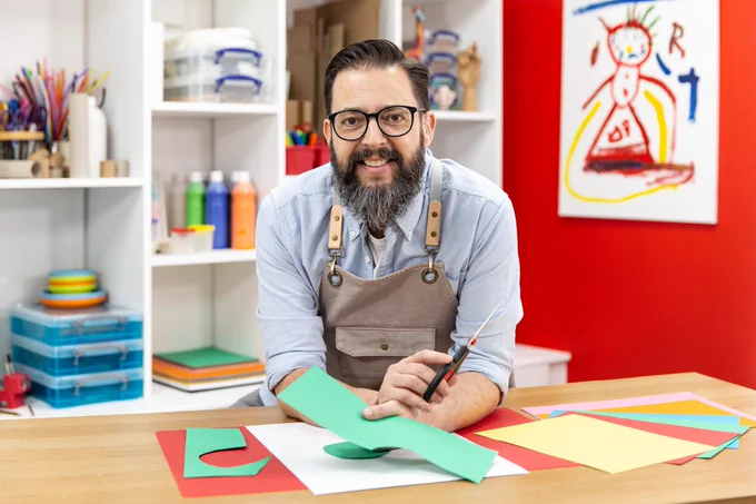 Art tools in the background, Christopher smiles, holding a large scissor and green paper. Wearing light blue shirt, brown work apron and thin black frame glasses. He has long beard, dark brown. He's kneeling his elbow on a table with colours paper spread out.