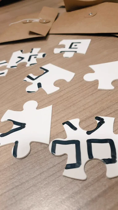Se ven una serie de piezas de puzzle en blanco con diferentes símbolos, que juntos formarían un código secreto. Detrás se ven dos sobres de papel craft donde se guardan estas piezas. 