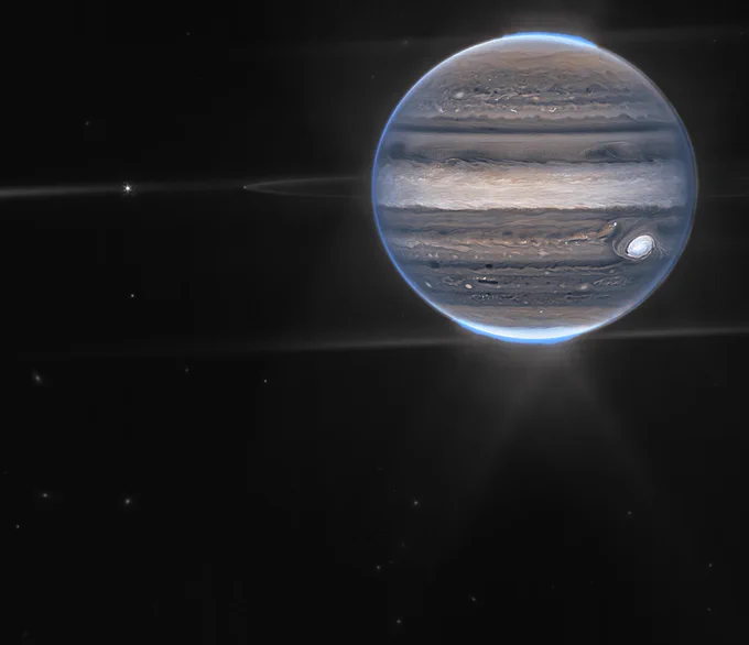 A wide field view showcases Jupiter in the upper right quadrant. The planet’s swirling horizontal stripes are rendered in blues, browns, and cream. Electric blue auroras glow above Jupiter’s north and south poles. A white glow emanates out from the auroras. Along the planet’s equator, rings glow in a faint white. These rings are one million times fainter than the planet itself! At the far left edge of the rings, a moon appears as a tiny white dot. Slightly further to the left, another moon glows with tiny white diffraction spikes. The rest of the image is the blackness of space, with faintly glowing white galaxies in the distance.

