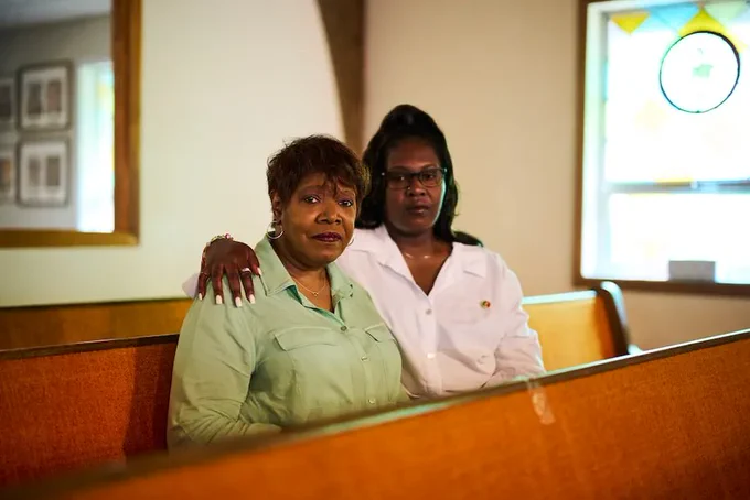 Two Black women sit together in a church pew, the younger woman's arm around the elder's shoulders. They have sad expressions on their faces.