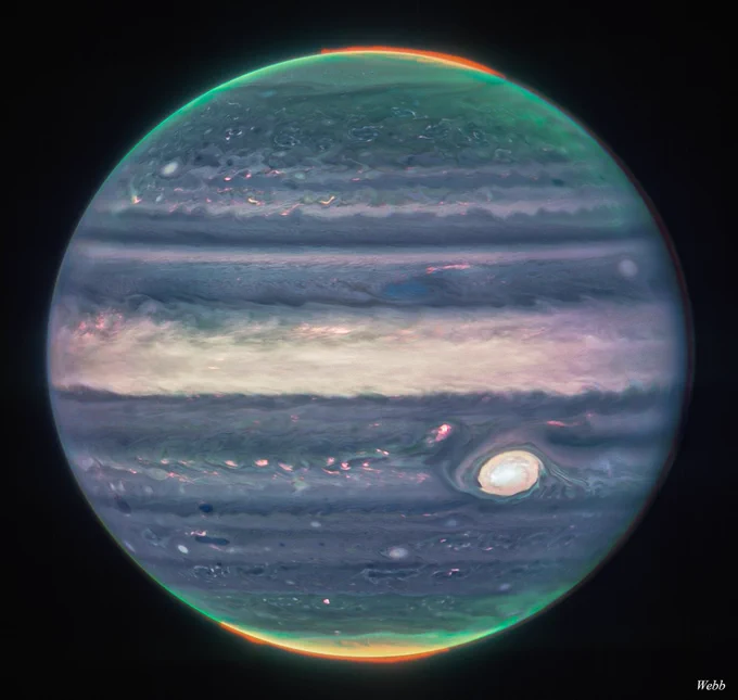 The planet Jupiter is seen in infrared light, appearing in cloudy tones of purple, beige, and pink. At its north and south poles, aurorae shine in bright green, red, and yellow hues.
