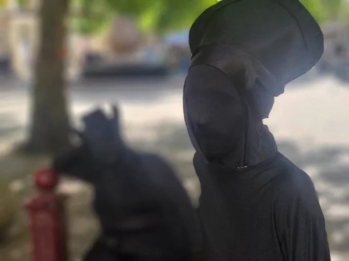 Foreground: Performer in black morph suit representing a chess Bishop looks at the camera in focus