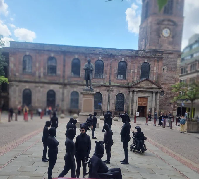 A large group of performers dressed in black morph suits representing chess pieces are strategically placed in St Ann's Square which is overlooked by a statue and Church with clock tower.