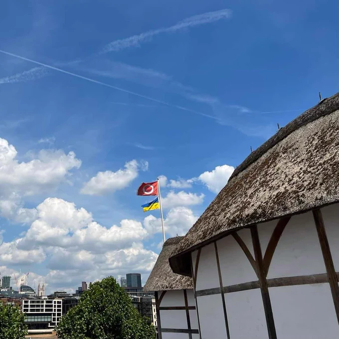 A photograph looking at the outside of the Globe Theatre and the River Thames. From the roof of the Globe, two flags fly and flutter in the breeze. One is red and shows the circular logo of Shakespeares Globe, the other is the blue and yellow flag of Ukraine. A bright blue sky shines above, with fluffy white clouds.