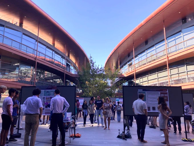Outdoor photo of researchers at large standing poster boards in a courtyard with the Clark Center building's many windows and red detailing in the background.