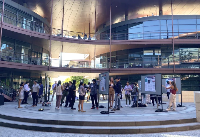 Outdoor photo of researchers at large standing poster boards in a courtyard with the Clark Center building's many windows and red detailing in the background.