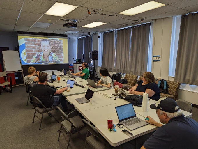 A group of Wikimedia editors sitting around a large table, watching a projector screen that is streaming the Wikimedian of the Year awards