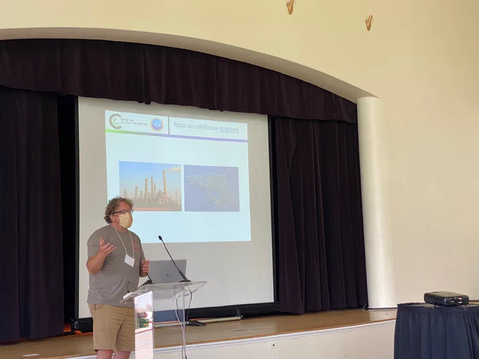 Professor standing in front of a slide with images of factories and cells to introduce how cells might be used for different applications