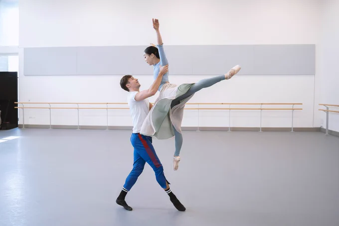 Joseph and Sarah are dancing in a rehearsal studio. Joseph has lifted Sarah off the ground, both of her hands are in the air and one of her legs is outstretched backwards. 