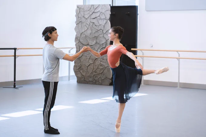 Kevin and Rachael are dancing in a rehearsal studio. Rachael is balancing on one leg with her other leg outstretched behind her. Kevin is stood in front of her, holding one of her hands.