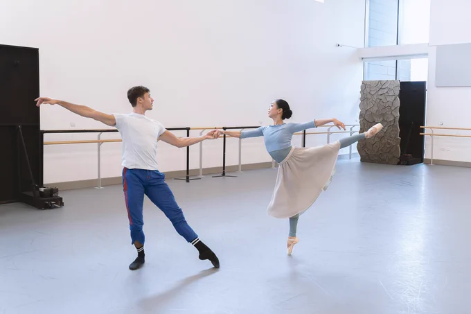 Joseph and Sarah are dancing in a rehearsal studio. Sarah is balancing on one leg with the other leg outstretched behind her. Joseph is stood next to her and is holding Sarah's hand.