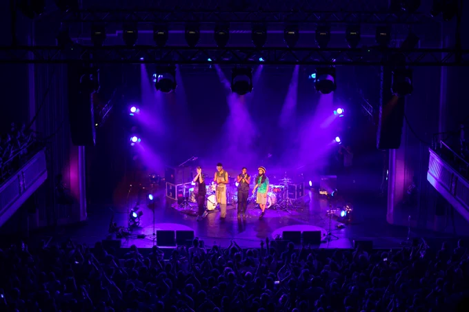 Four men stand smiling in front of two drum sets on
stage in front of a full crowd of people applauding.