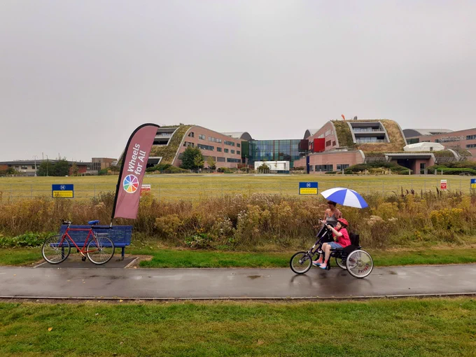 A young woman on a handcycle, with Alder Hey Children's Hospital in the background. Her mother shelters her from the rain with an umbrella. To the left of frame is a 2-wheel bicycle, with a Wheels for All flag attached to the set post