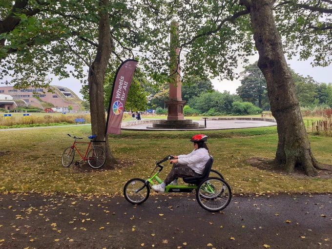 A young woman riding a Tomcat Dragon semi-recumbent cycle in Springfield Park, on a path through trees. In the background is a large monument. Next to a tree is a 2-wheel bicycle with a Wheels for All flag attached to the seat post with a bracket 