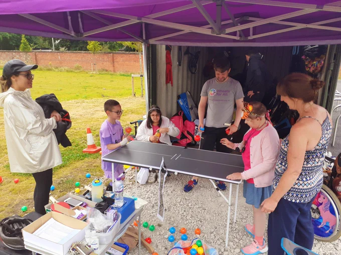 Two children play inclusive table tennis beneath a purple Wheels for All gazebo, while mums and Wheels for All staff look on