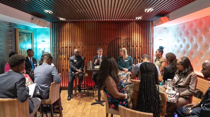 Secretary Blinken sits at a table at the front of a restaurant and addresses a group of young people.