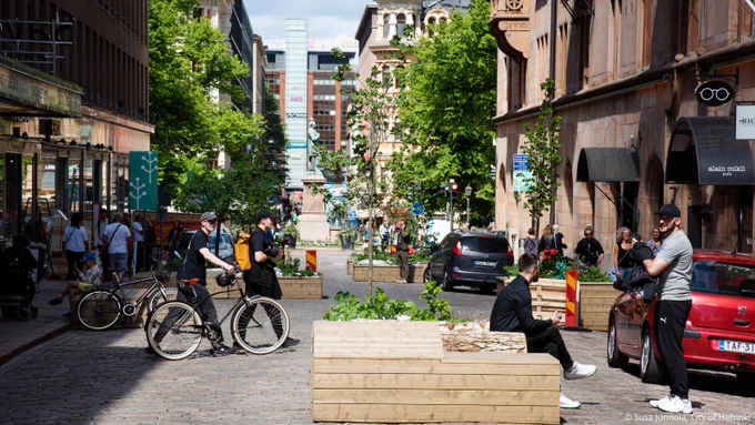 Photo of the wooden seating installed on Kasarmikatu street in Helsinki, Finland. People are using the space by sitting on the temporary wooden seats, as well as walking. 