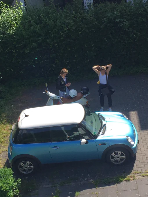 Ein Mini Cooper in mittel blau metallic mit weißem Dach, dahinter zwei Mädchen mit einem hellblauen  Vespa Motorroller
