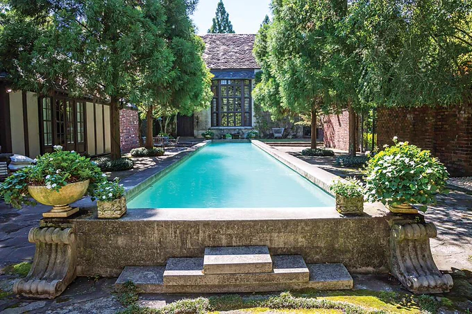 In the backyard of a Tudor-style house, an in-ground pool is surrounded by an ornate concrete border, plants potted in urns, and shady trees.