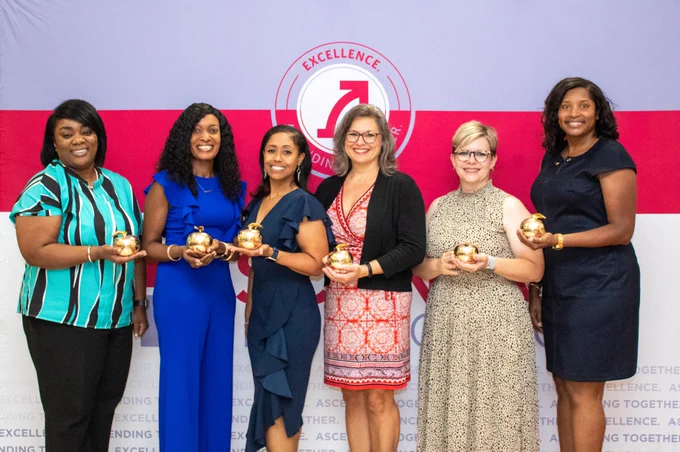Six women stand side by side holding a golden apple.