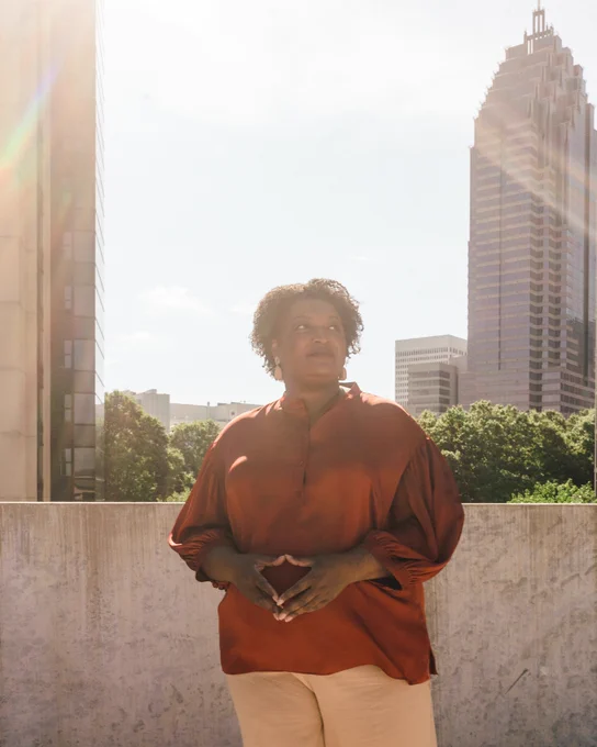 A photo of Stacey Abrams standing and looking up. Behind her is a skyscraper building in Atlanta.