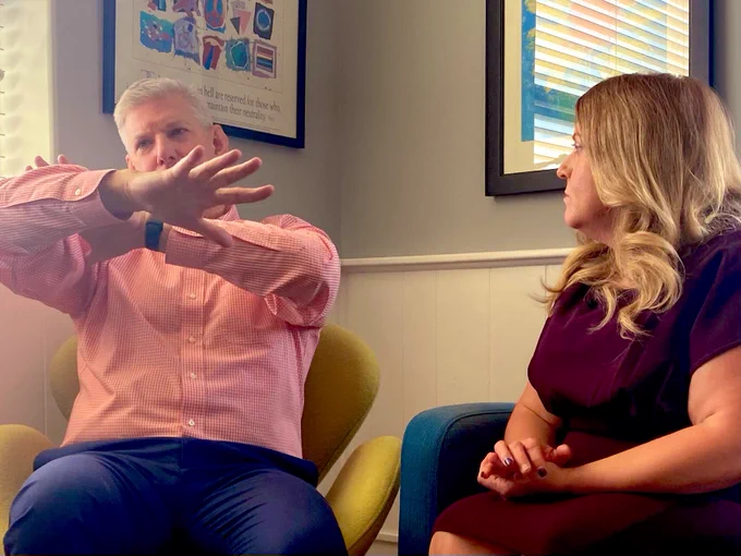 Craig and Anne on interview set, Craig gesturing with arms in an X position, Anne looking on with a “what is he saying?” expression