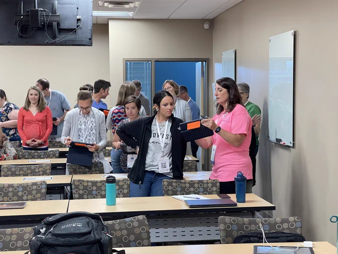 Teachers in a classroom. They are standing and engaging with each other. Some are showing each other their iPad screens as they engage in learning.
