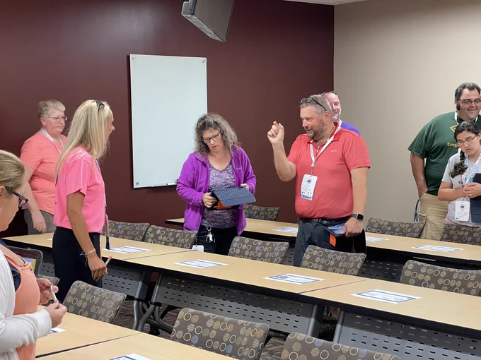 Teachers in a classroom. They are standing and engaging with each other. Some are showing each other their iPad screens as they engage in learning.