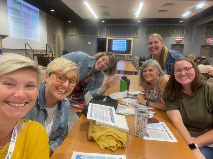 6 Teachers work at a long table in a presentation room.  They are posing for the group picture!