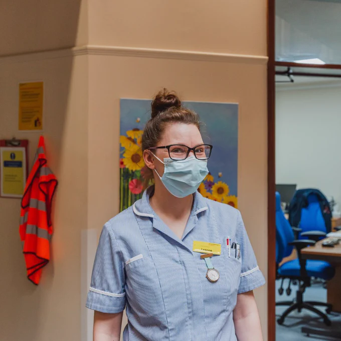 Nurse wearing uniform, mask and glasses, looking at the camera.