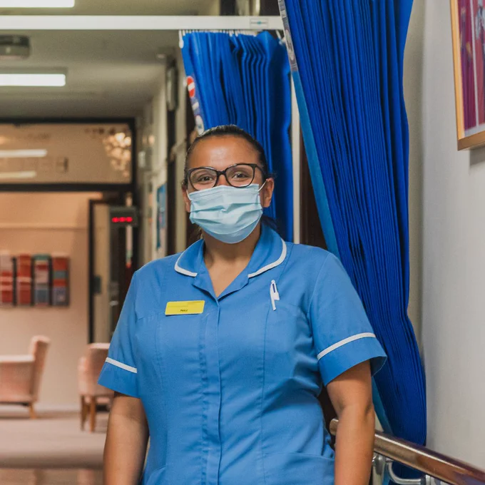 Nurse wearing uniform, mask and glasses, looking at the camera in the ward corridor.