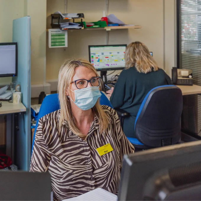 Rose, wearing glasses, mask and a zebra print top, working at a computer in an office.