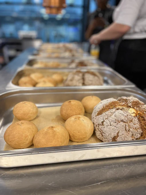 Five pans of bread in a row on an industrial table. Each pan has eight white bread rolls and a loaf of soda bread dusted with flour