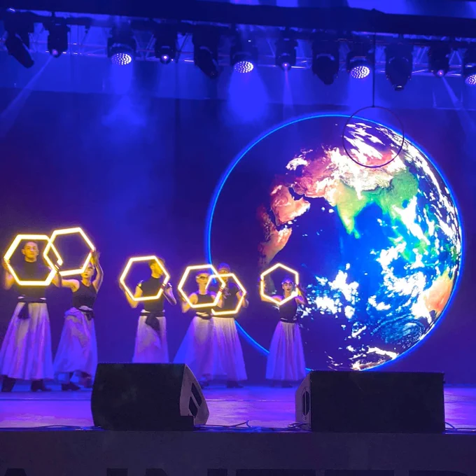 Dancers on stage holding glowing hexagons. A large Earth image is shown in the background.